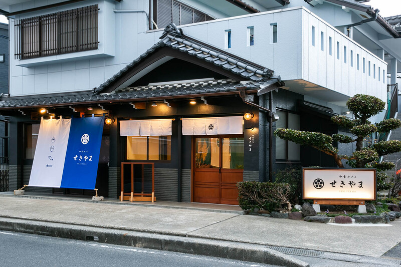 外観写真：以前から残る破風の黒しっくいに合わせて全体の色調を統一。既存の木枠を利用した屋外サイン等で仕出し屋からの名残を残しています。内観写真：ギャラリー用の可動壁等により客席（個人、団体）、コミュニティースペース等を緩やかに仕切り落ち着きのある空間にしています。